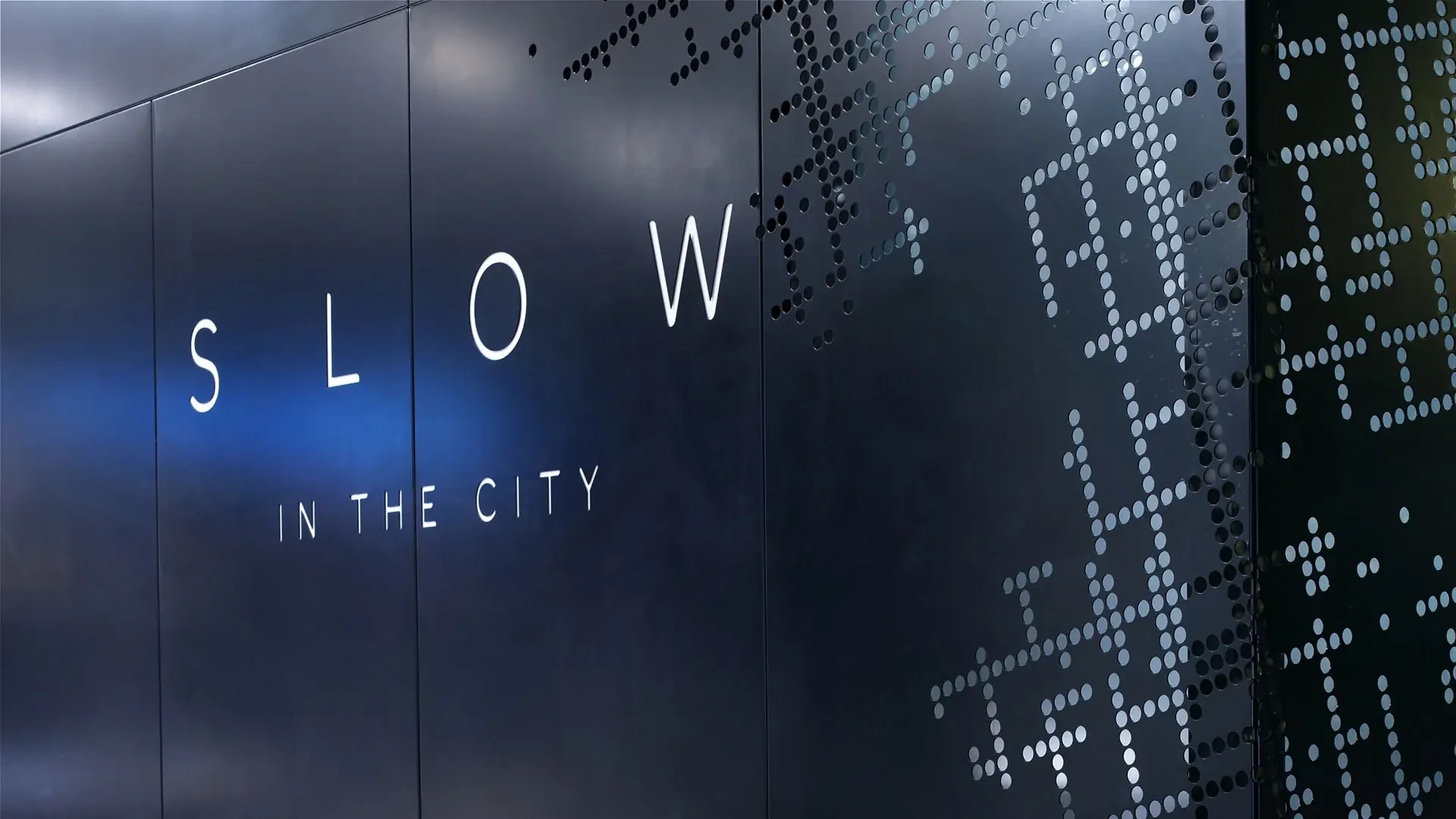SLOW in the City wordmark set against a dark perforated wall inspired by a city street grid