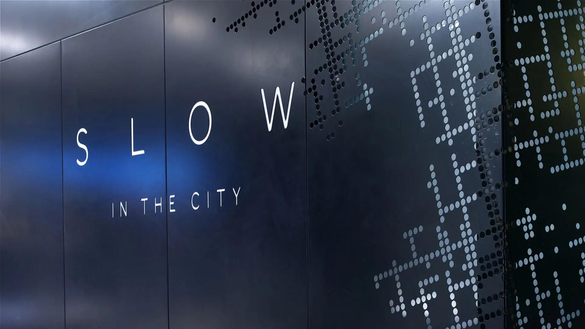 SLOW in the City wordmark set against a dark perforated wall inspired by a city street grid